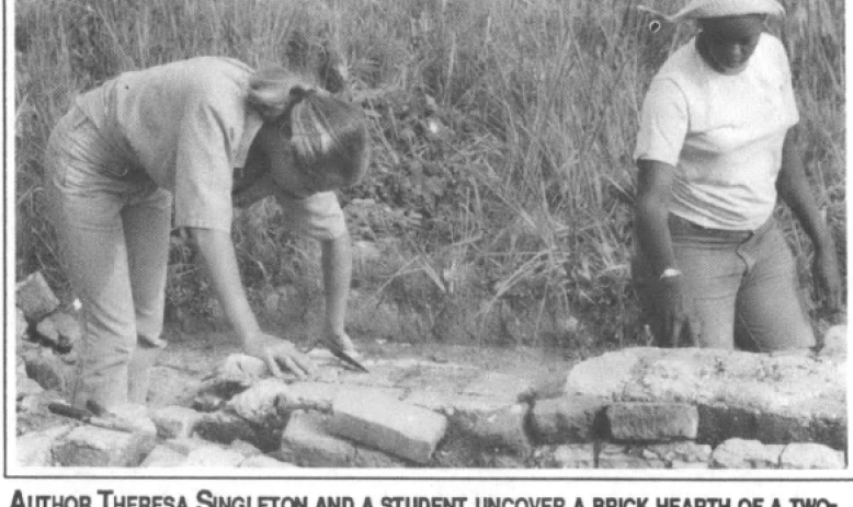 two people in a field, kneeling on dirt with text "AUTHOR THERESA SINGLETON AND A STUDENT UNCOVER A BRICK HEARTH OF A TWO FAMILY SLAVE DWELLING ON BUTLER ISLAND, A FORMER PLANTATION IN GEORGIA."
