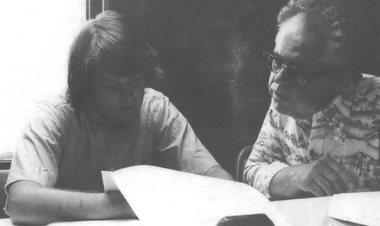 Black and white photo of two white men, one younger and one older, looking through papers at a table