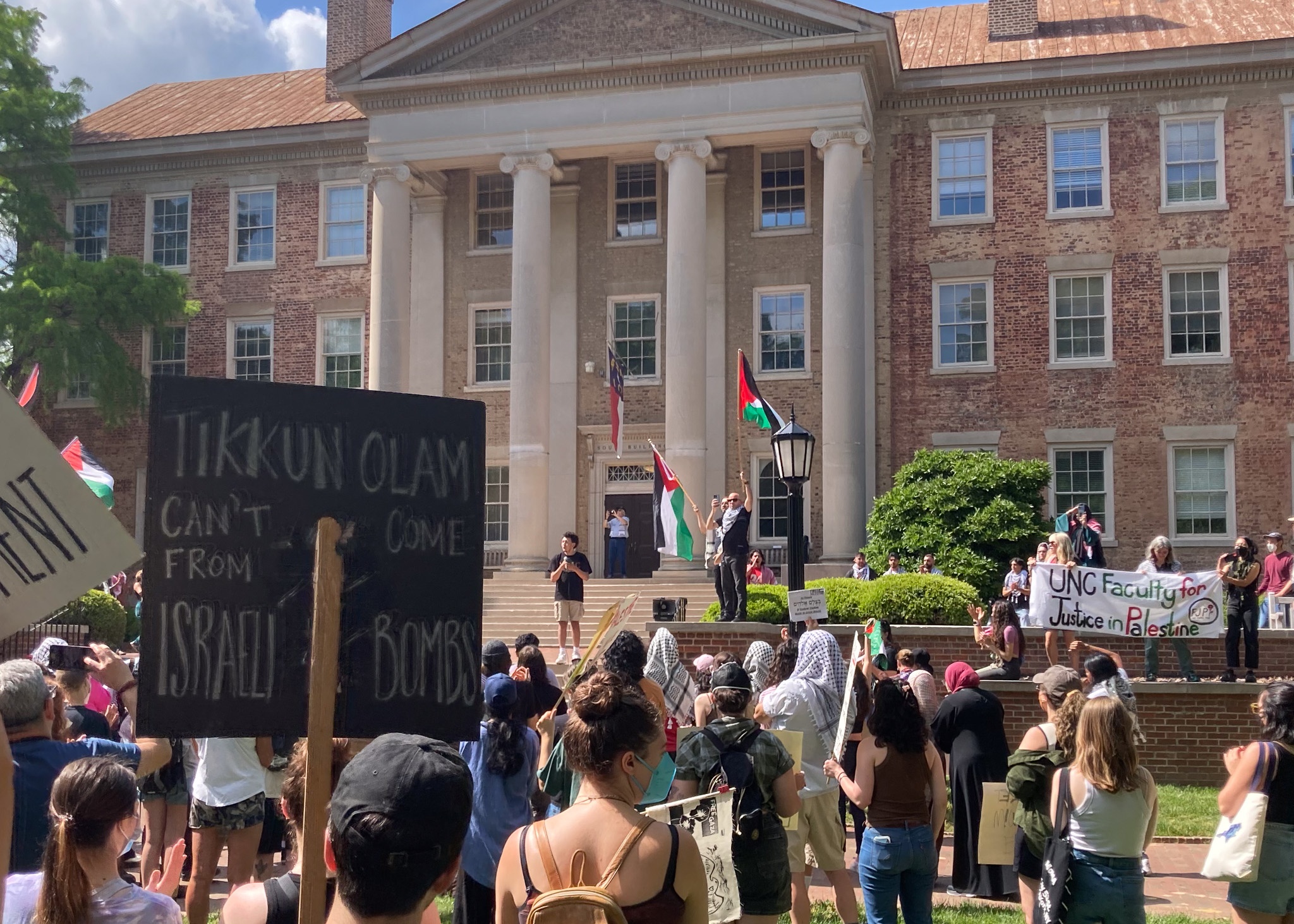 From-behind image of protestors in front of UNC's Wilson library. Two signs are visible: "Tikkun Olam can't come from Israel's bombs" and "UNC Faculty for Justice in Palestine." Speakers at the front hold Palestinian flags.