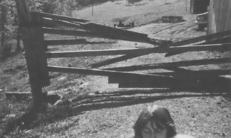 Black and white photo of girl with animal on her shoulder in front of falling-down fence