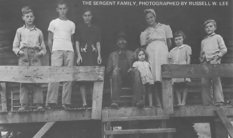 Black and white photo of white family, roughly 8 people, posed on wooden porch