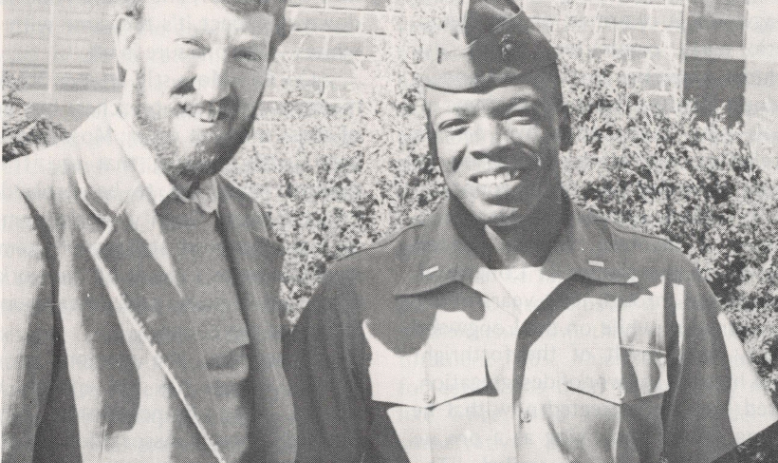 Black and white photo of two young men smiling at the camera, one white and one Black, dressed in military uniform