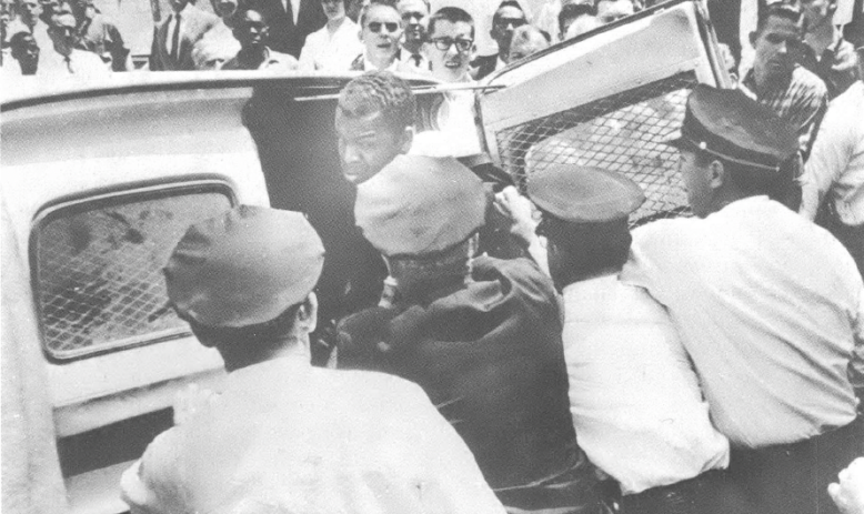 Black and white photo of people being pushed into car with mob around