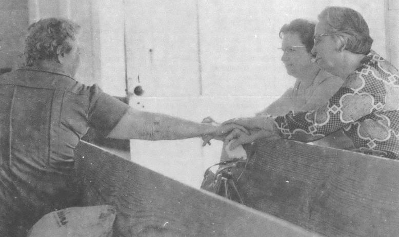 Black and white photo of three white women sitting in church pews, one woman shaking another woman's hand