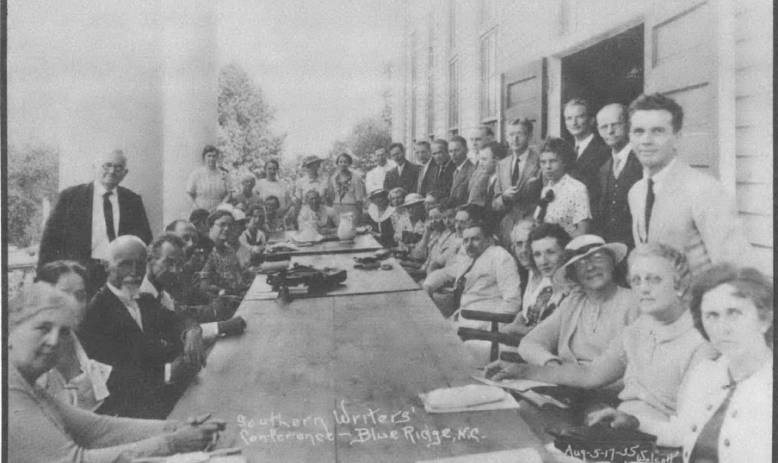 Black and white photo of a couple dozen people gathered around a table, facing the camera