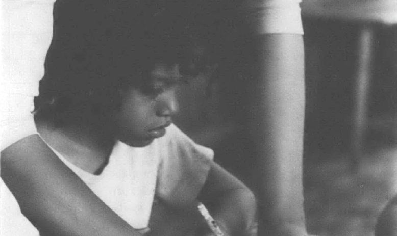 Black and white photo of Black woman helping Black child, seated at desk, write