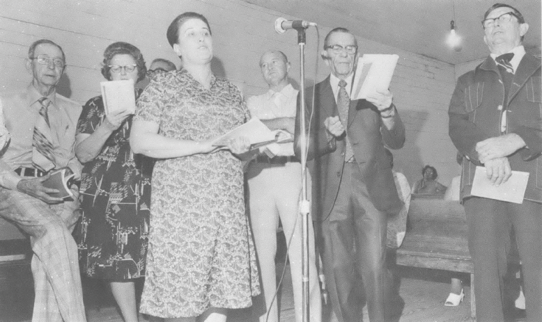 Black and white photo of older white people singing around a microphone in a church