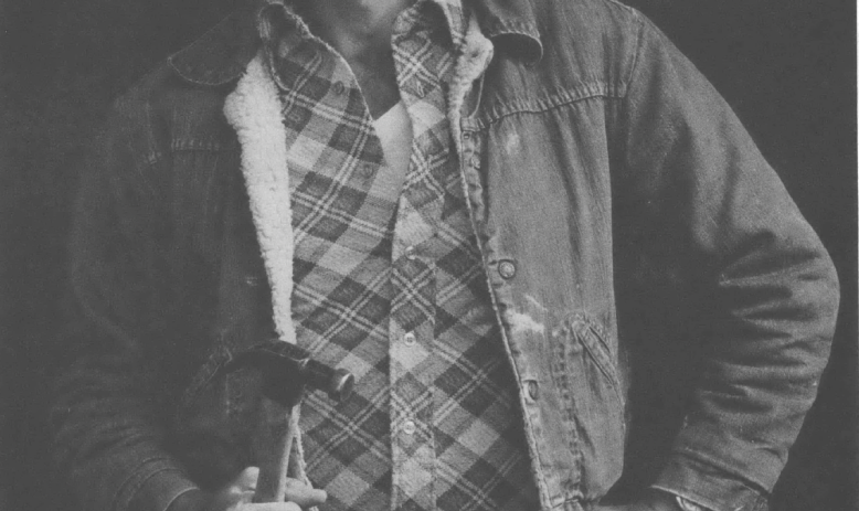 Black and white photo of young man wearing plaid shirt, jacket, and tool belt