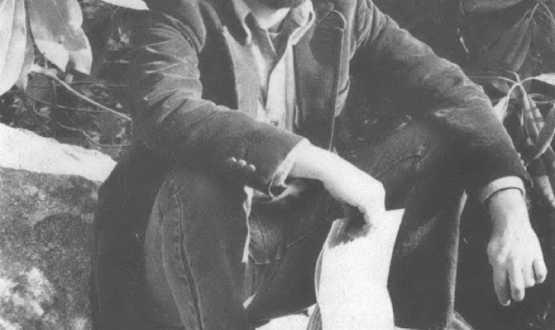 Black and white photo of young man wearing trucker hat holding papers sitting on the ground