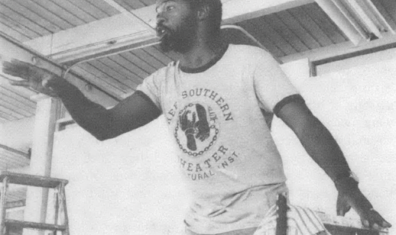 Black and white photo of Black man in t-shirt performing on stage, pointing at audience