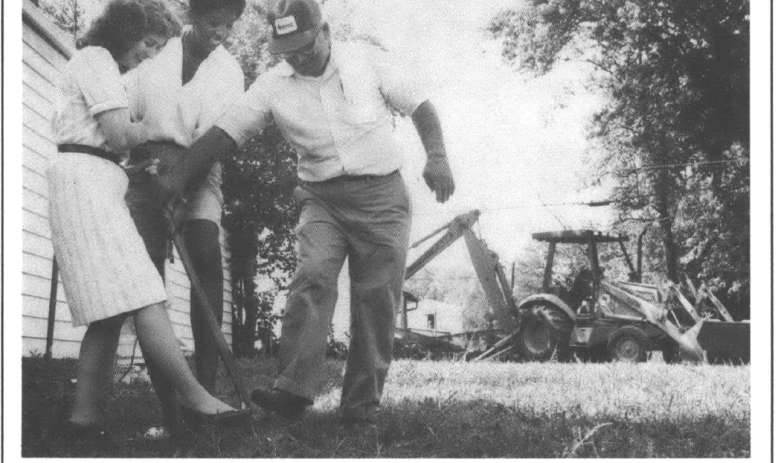 three people breaking ground with a single shovel on a peice of land boaldoazer and houses in the background 