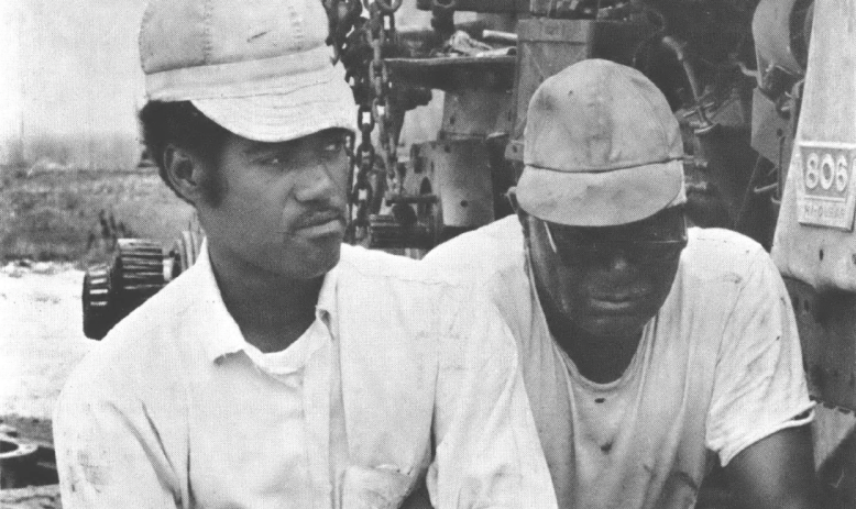 Black and white photo of two Black men, one younger and one older, both wearing trucker hats and standing next to a piece of agricultural equipment