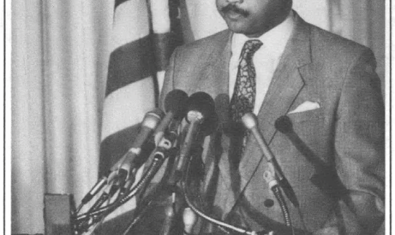 Man in a suit with glasses standing at a podium with 6 mics giving a press conference in front of a national seal and american flag