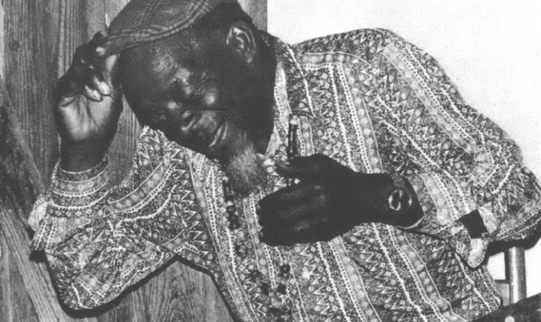 Black and white photo at an angle of Black man wearing patterned button-down shirt and cap, appears to be singing