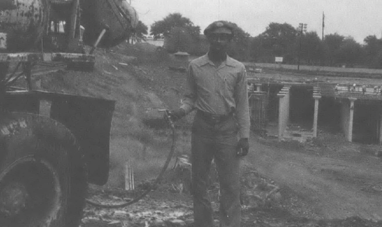 Black and white photo of Black man holding hose next to what looks like a cement mixer