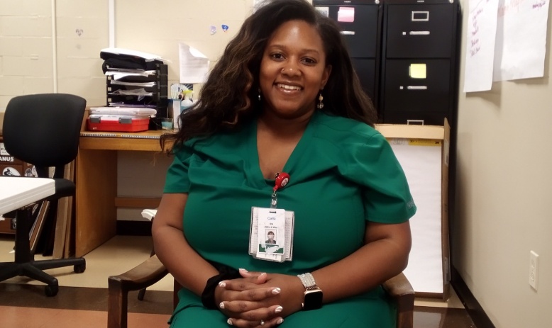 Black woman with long hair wearing green scrubs, sitting in chair and smiling