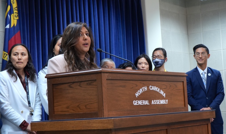 An Indian American woman speaks at a wooden podium that has the words "North Carolina General Assembly" attached to the front. Several other people who are Asian American stand behind the woman, watching her speak.