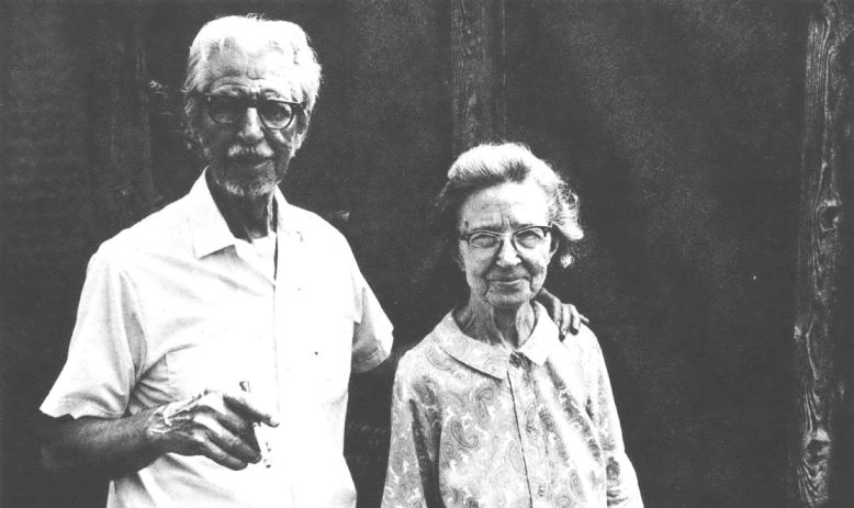 Black and white photo of an elderly white couple, the white-haired, balding man on the left wearing glasses and a collared shirt, and the shorter woman on the right in glasses and a dress.