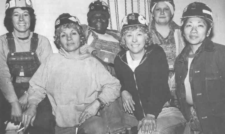 Black and white photo of six women of different races and ethnicities in coalmining gear