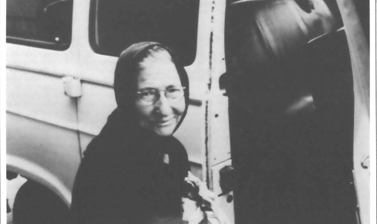 Photo of older woman with scarf around her head next to a car