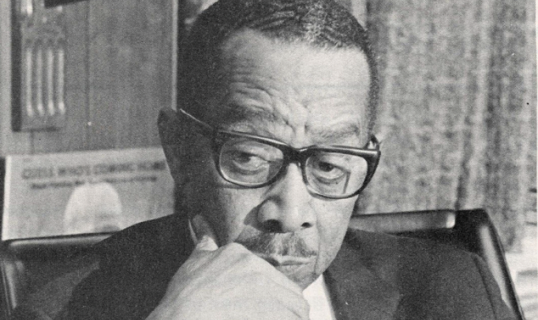 Black and white portrait of Black man with glasses, in suit, and hand on chin posed in front of bookcase