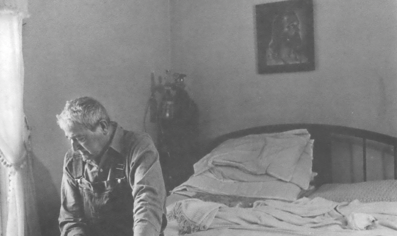 Black and white photo of elderly white man sitting at the end of a bed looking down at the floor