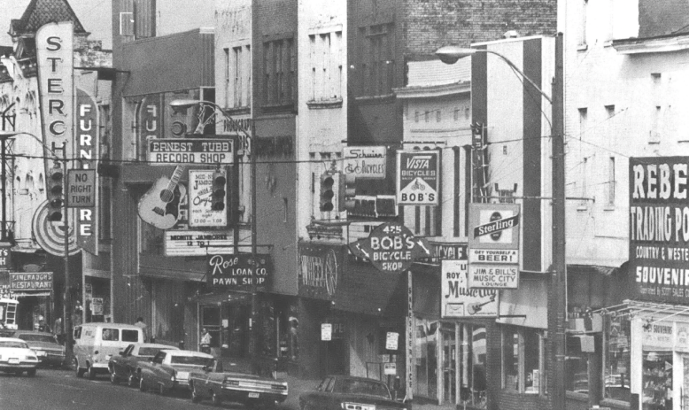 Black and white photo of city street with several record and music shops, and cars parked on the street in front