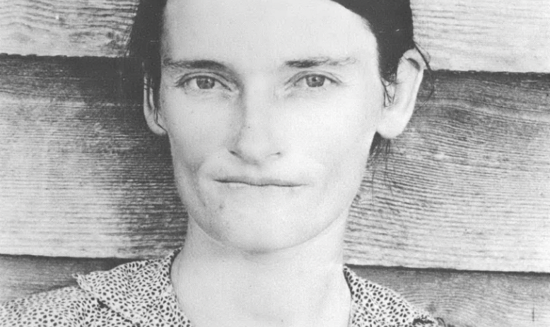 Black and white portrait of young white woman with hair pulled back standing against wood paneled building