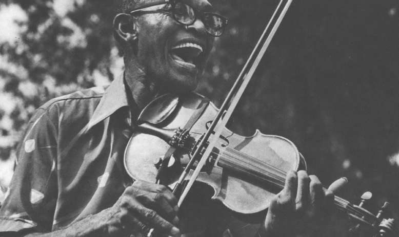 Black and white photo of Black man in glasses and collared shirt playing fiddle and laughing