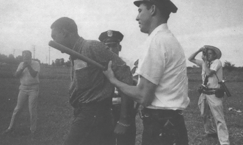 Black and white landscape photo of cops arresting a Black man in a field, with photographers behind