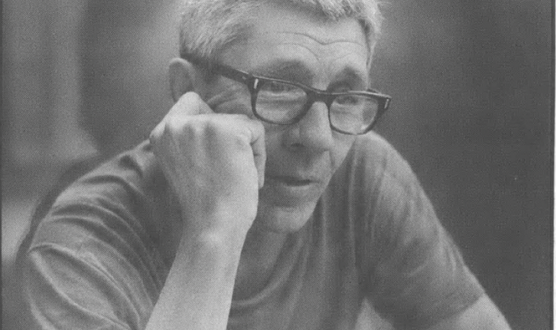 Black and white image of white man with grey/white hair and glasses, head resting on one hand