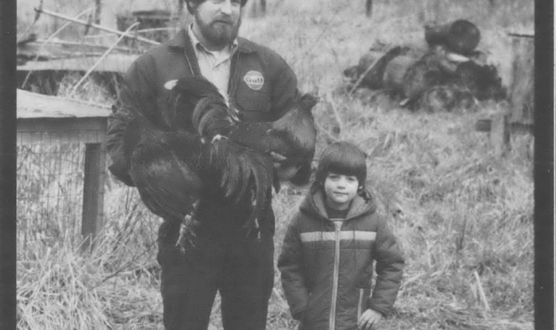 Black and white photo of white man and child standing outside against farm backdrop, wearing coats