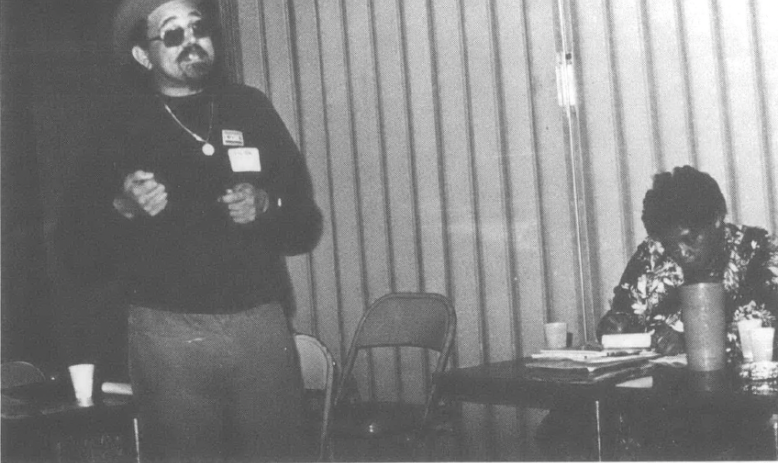 Photo of Black man in cap and sunglasses standing with mic speaking, Black woman at a table in the background. Caption reads Ron Chisom at Gulf Coast tenant workshop
