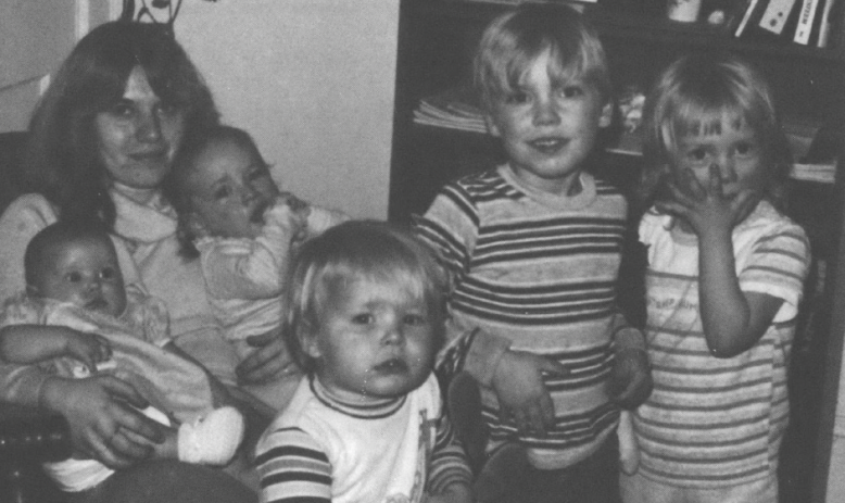 Black and white photo of seated white woman holding two babies with three other young children standing in front of her