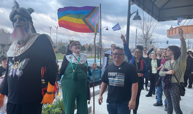 The image depicts a group of individuals smiling, holding gay and trans pride flags, and walking along a sidewalk in a parade on Trans Day of Visibility.