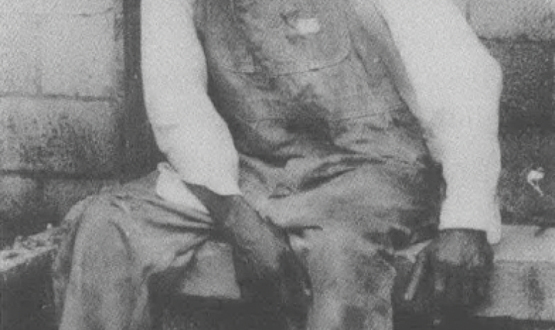 Black and white photo of older Black man in overalls and hat sitting on a bench outside of a building, slightly hunched over