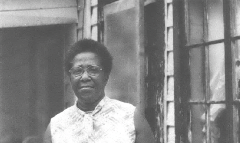 Black and white photo of middle-aged Black woman standing in front of a home