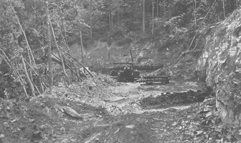 Black and white photo of hillside being excavated, with trees on their sides and forest surrounding