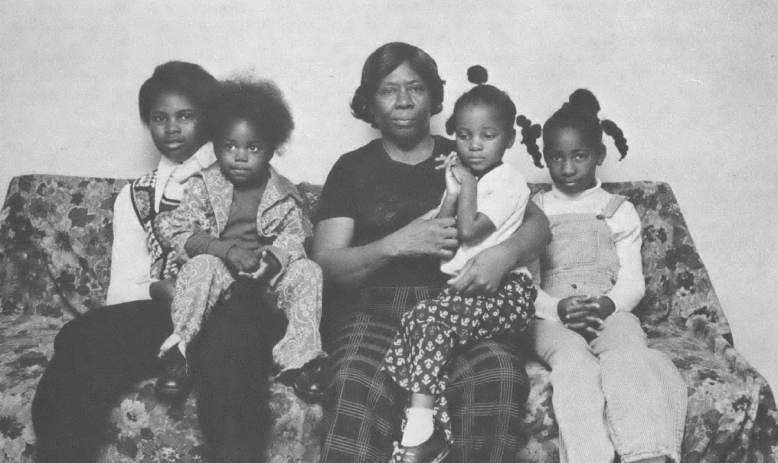 Black and white family photo of Black woman with four young children sitting on a couch inside a home. 