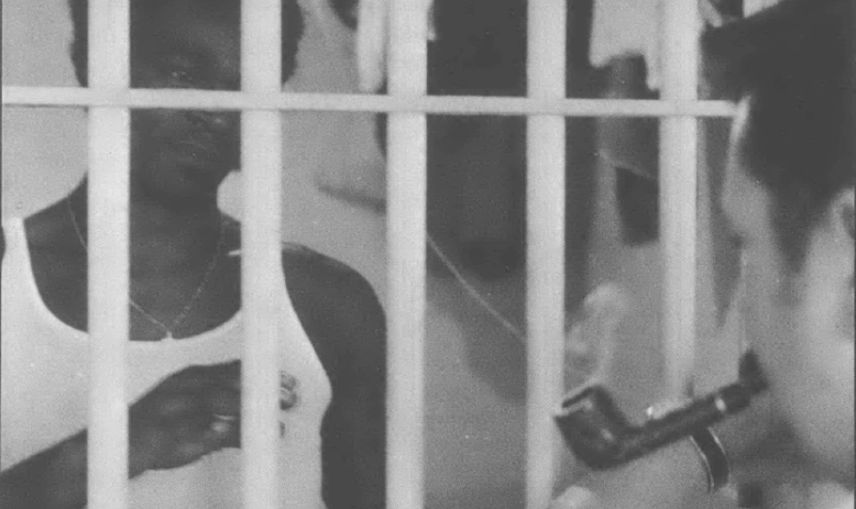 Black and white photo of a white man smoking a pipe facing a Black man who is in a cell