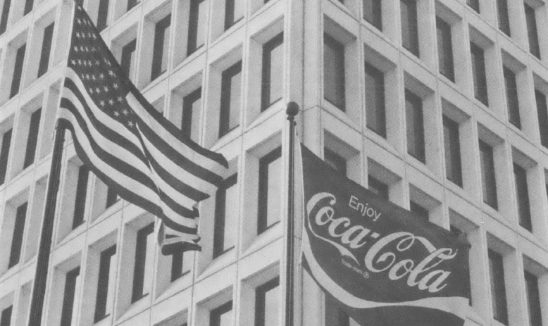 Black and white photo of American flag and Coca-Cola flag flying in front of high-rise building