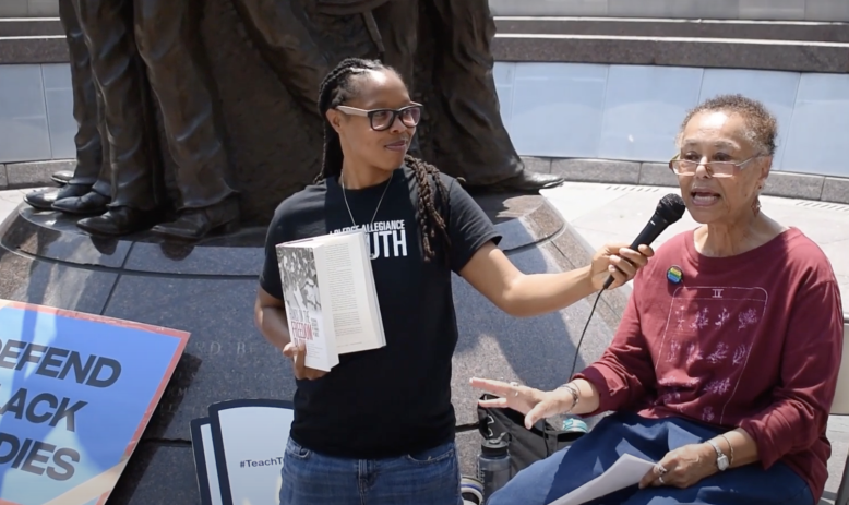 A Black woman wearing a black t-shirt holds out a microphone for another older Black woman who is seated and wearing a red t-shirt to speak into. They are at the African American Civil War Memorial in Washington, D.C. A sign that reads "Defend Black Studies" lays in the corner.