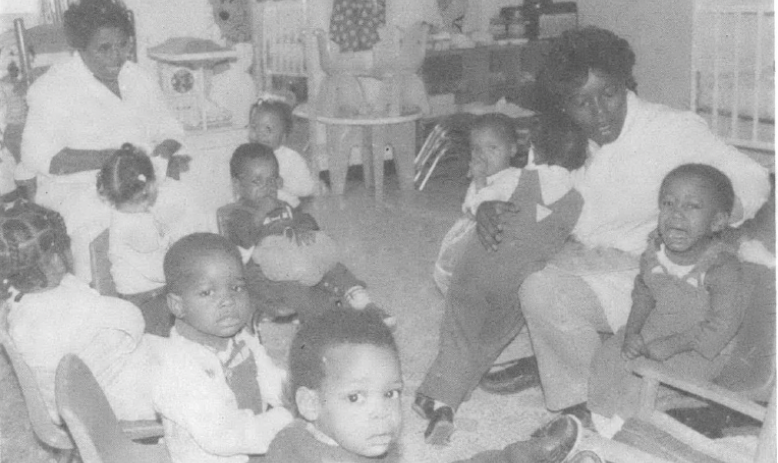Two teachers and several children sit on the floor of a daycare center