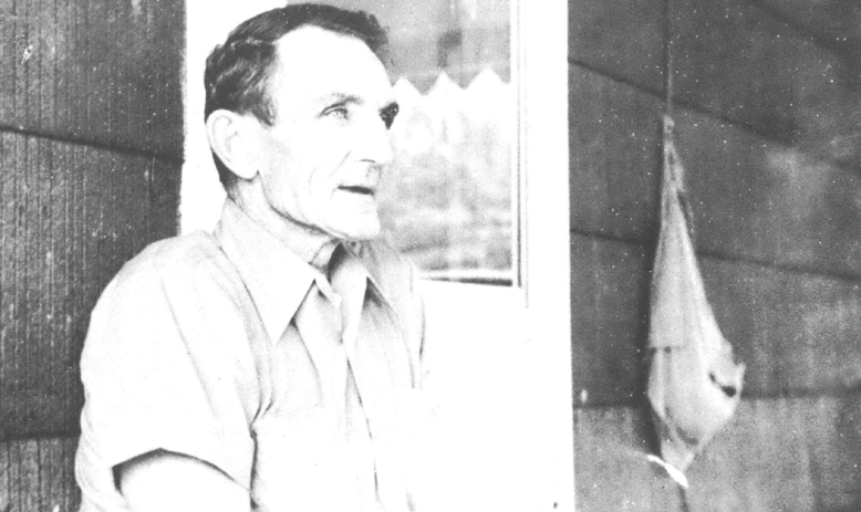 Black and white photo of older white man seated outside in front of the window of a house, talking and looking at someone away from the camera