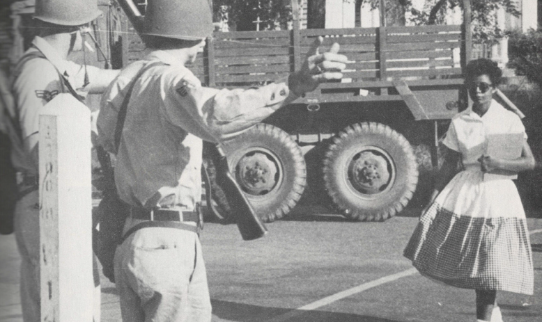 Black and white photo of young Black student in dress and sunglasses walking towards two National Guard members