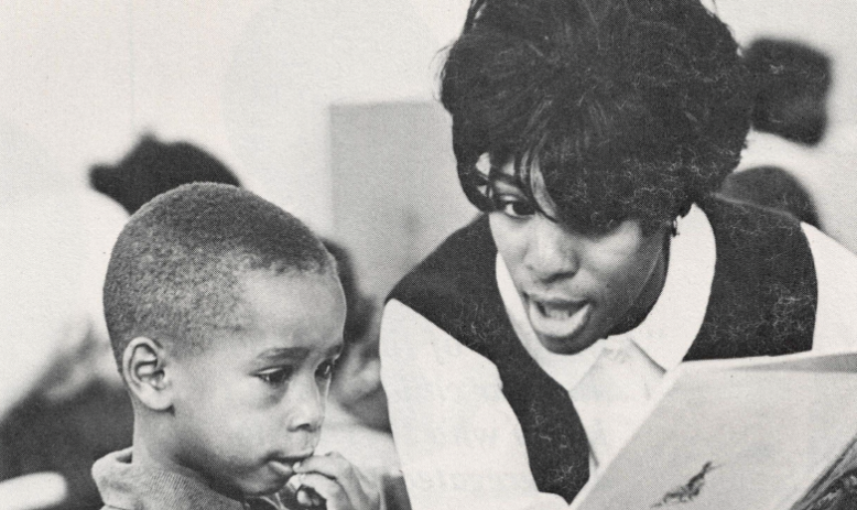 Black and white photo of Black teacher explaining a paper to young Black student in classroom