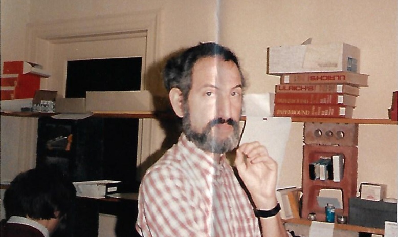 Scanned photo with crease through the middle showing man standing in an office with his hand to his chin, wearing a red and white checked shirt