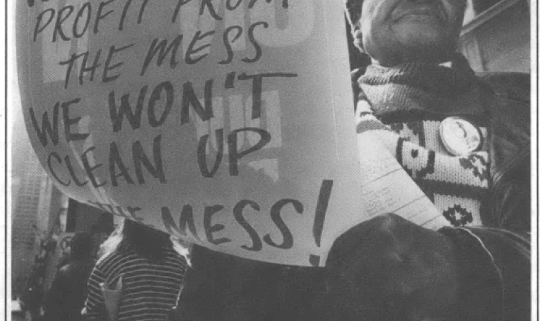 Woman in a scarf, pin, and coat in the streets at a protest with a sign that reads "WE DIDN'T make the mess WE DIDN'T profit from the mess WE WONT CLEAN UP THE MESS!"