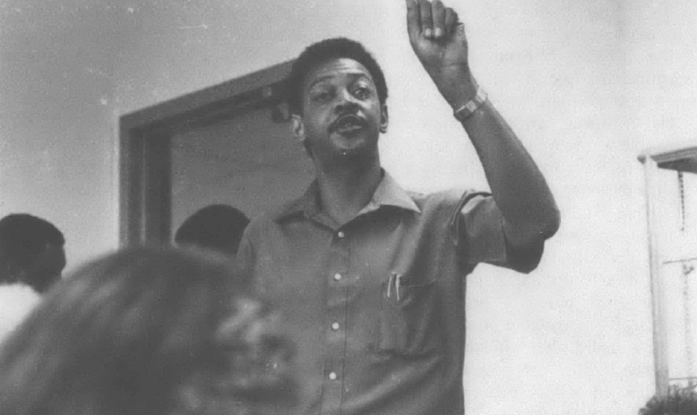 Black and white photo of Black man speaking to group of people in a room
