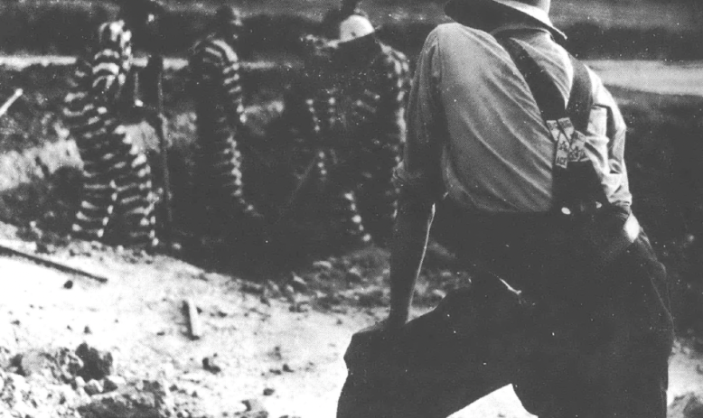Black and white photo of a man with his back turned to the camera watching prisoners in black and white jumpsuits digging a hole.